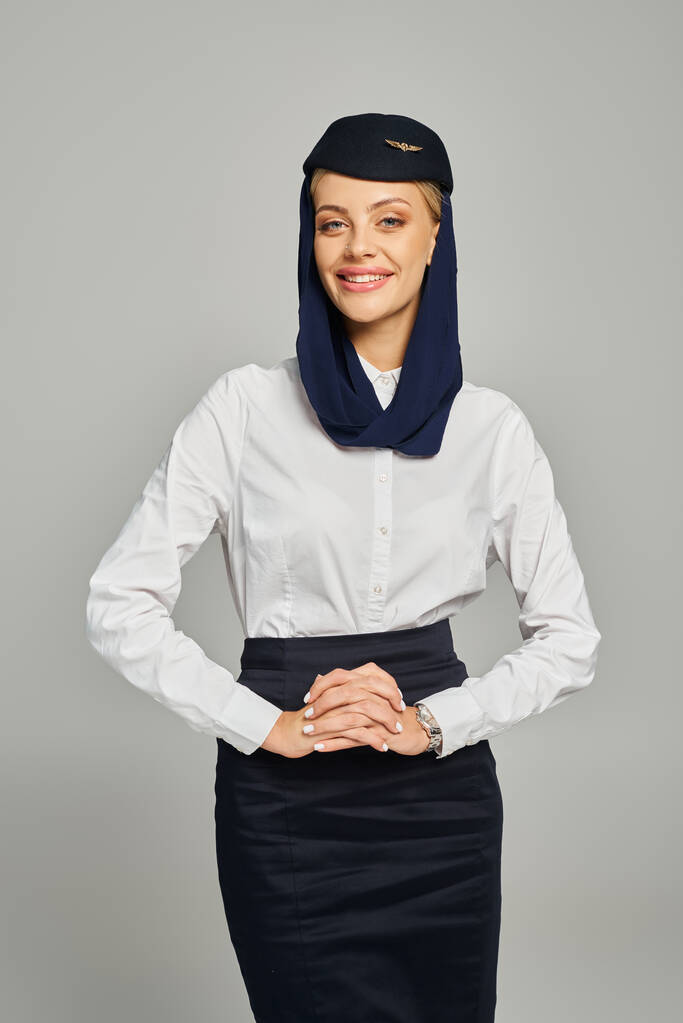 χαμογελαστή αεροσυνοδός της αραβικής αεροπορικές εταιρείες με μαντίλα και λευκή μπλούζα χαμογελώντας στην κάμερα σε γκρι - Φωτογραφία, εικόνα