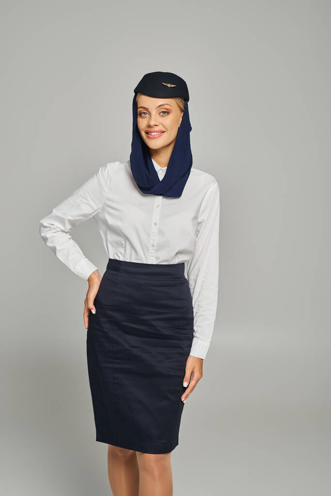 stijlvolle en vrolijke stewardess in Arabische luchtvaartmaatschappijen uniform poseren met de hand op de heup op grijs - Foto, afbeelding