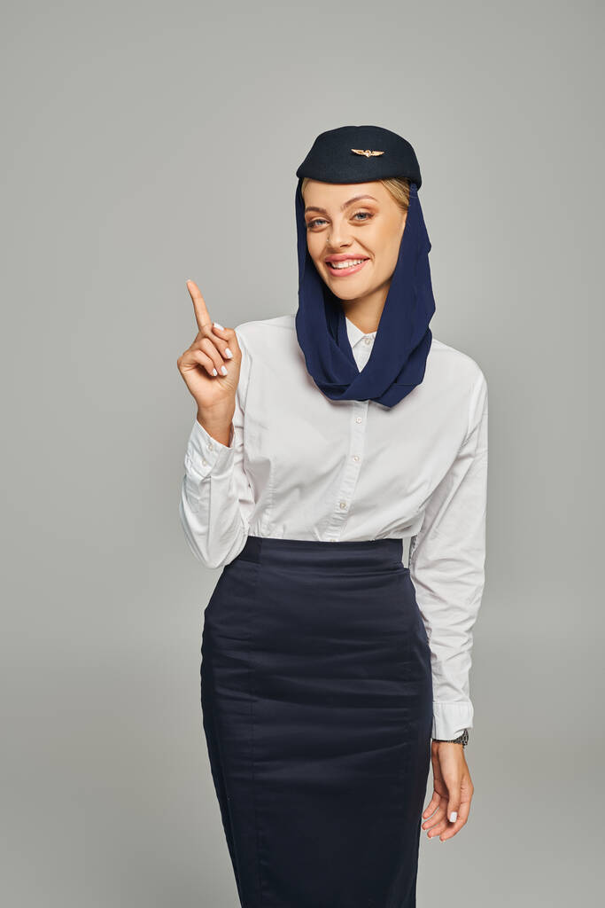 Genç Arap havayolları hostesi başörtüsü ve üniforması ile gri arka planda parmakla işaret ediyor. - Fotoğraf, Görsel