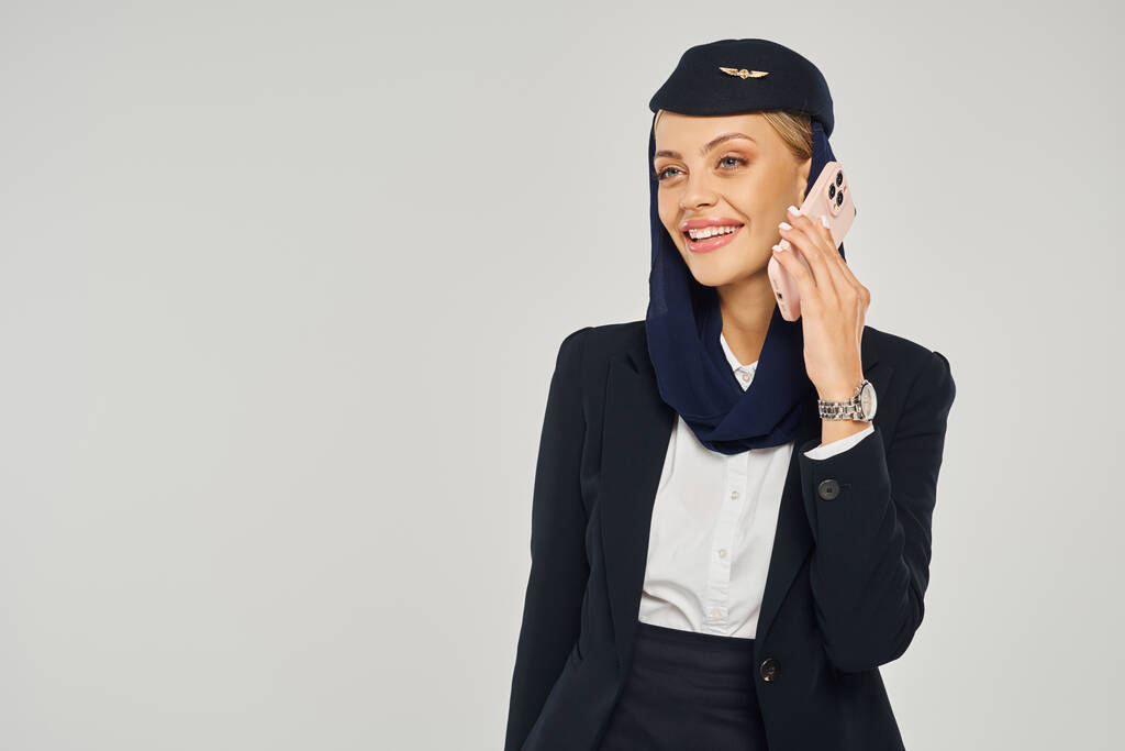 νέα και κομψή αεροσυνοδός σε αραβικές αεροπορικές εταιρείες μιλώντας στο κινητό τηλέφωνο σε γκρι φόντο - Φωτογραφία, εικόνα