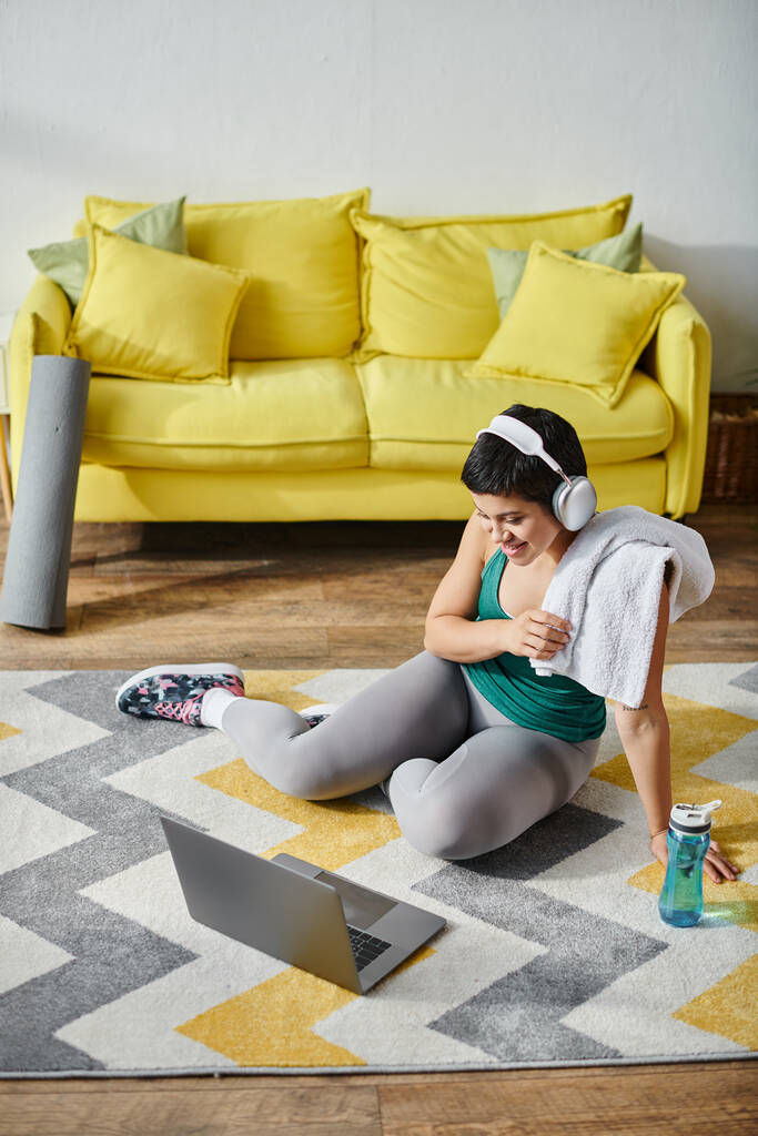 colpo verticale di donna gioiosa seduta sul pavimento con asciugamano accanto al computer portatile e bottiglia d'acqua, fitness - Foto, immagini