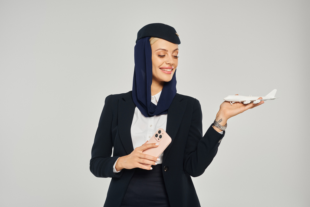 azafata sonriente de las aerolíneas árabes con teléfono móvil mirando el modelo de avión en el fondo gris - Foto, imagen