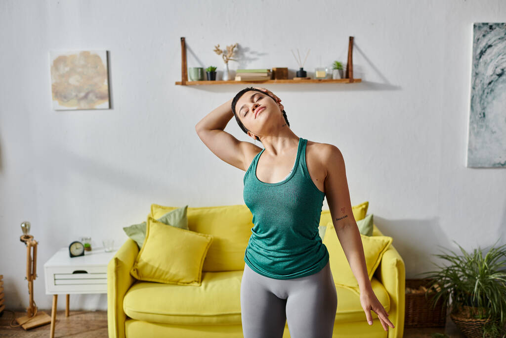 dobrze wyglądająca młoda kobieta z krótkimi włosami rozciągająca swoje ciało przed żółtą sofą, fitness - Zdjęcie, obraz