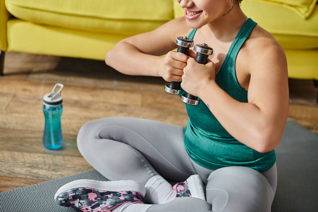 oříznutý pohled na smajlíka mladá žena sedící na podlaze cvičení s činkami, fitness a sport - Fotografie, Obrázek