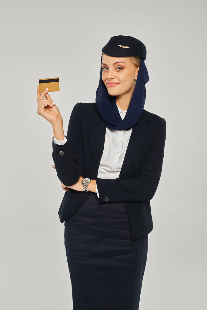 fröhliche arabische Stewardess in Uniform und Kopftuch mit Kreditkarte vor grauem Hintergrund - Foto, Bild