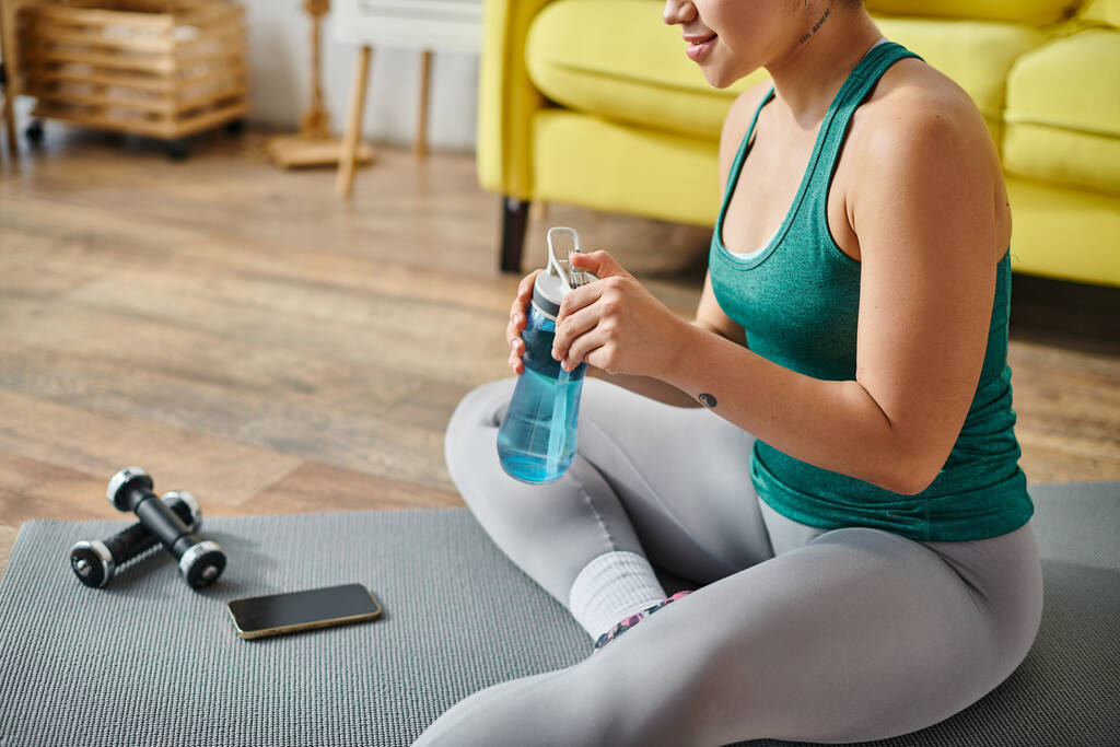 cropped view of young woman sitting on floor near dumbbells and phone with water bottle in hands - Photo, Image