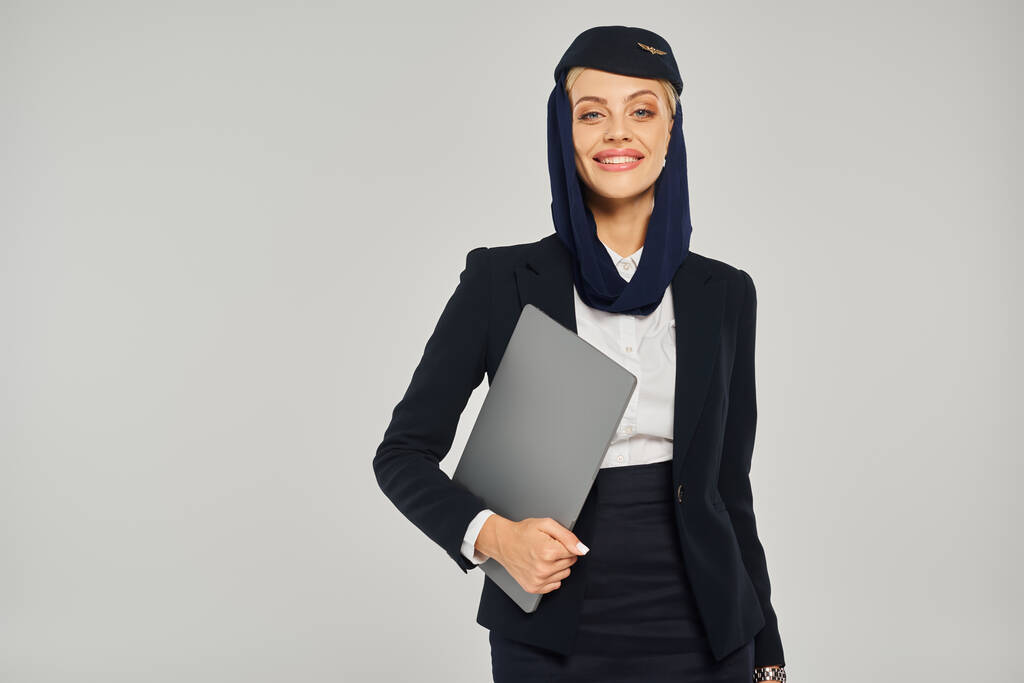 hostess di compagnie aeree arabe positive in uniforme con laptop che sorride alla macchina fotografica su sfondo grigio - Foto, immagini