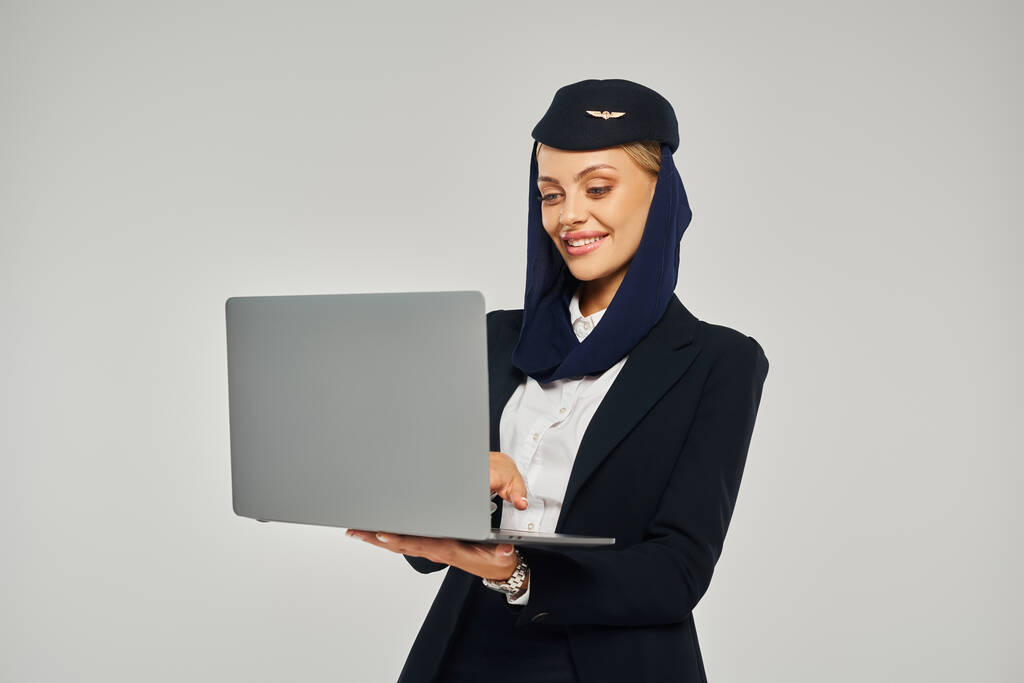 heureuse hôtesse de l'air arabe compagnies aériennes en uniforme d'entreprise à l'aide d'un ordinateur portable tout en se tenant sur gris - Photo, image