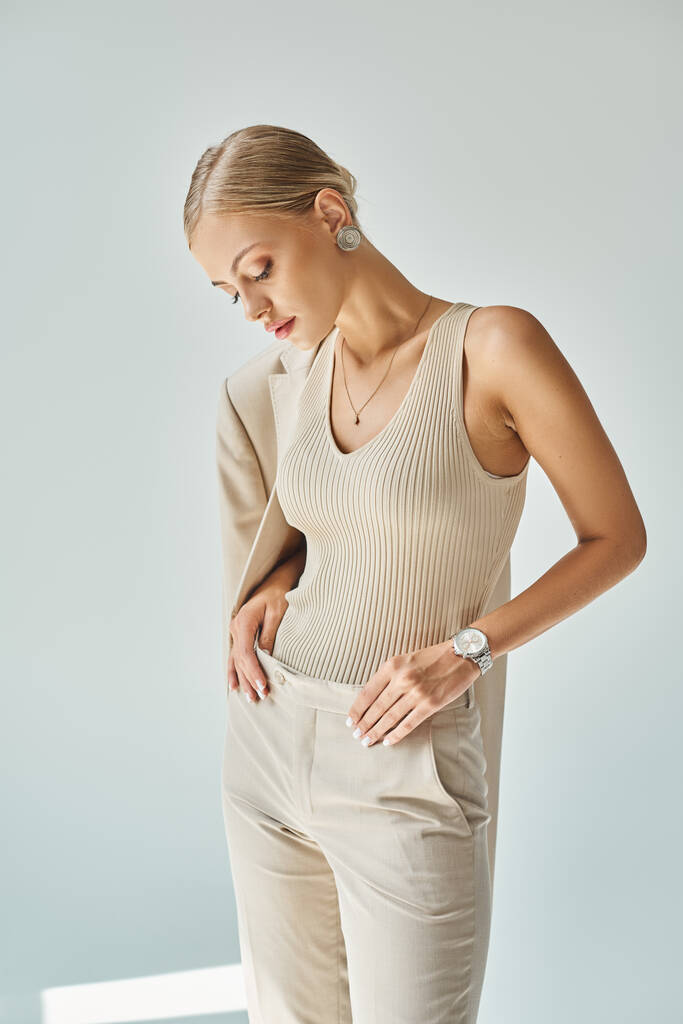 fashionable blonde woman in total beige clothes touching pants while standing on grey backdrop - Photo, Image