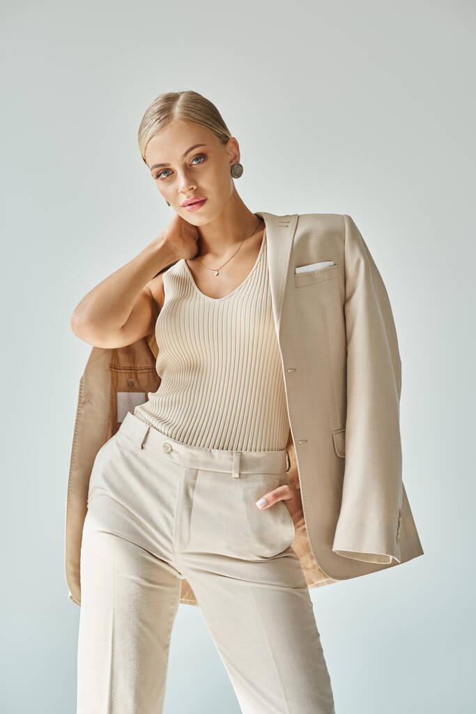 giovane modella in eleganti abiti beige toccando il collo e posando con mano in tasca su grigio - Foto, immagini