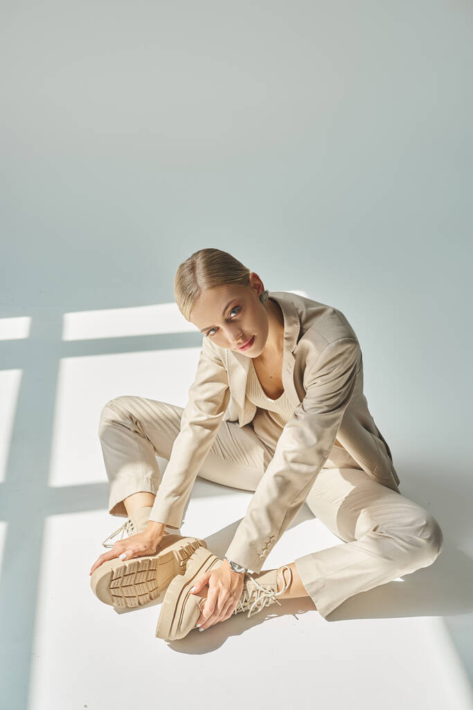 σαγηνευτική γυναίκα σε παστέλ ρούχα αγγίζοντας δεμένο μπότες, ενώ κάθεται στη λιακάδα σε γκρι φόντο - Φωτογραφία, εικόνα