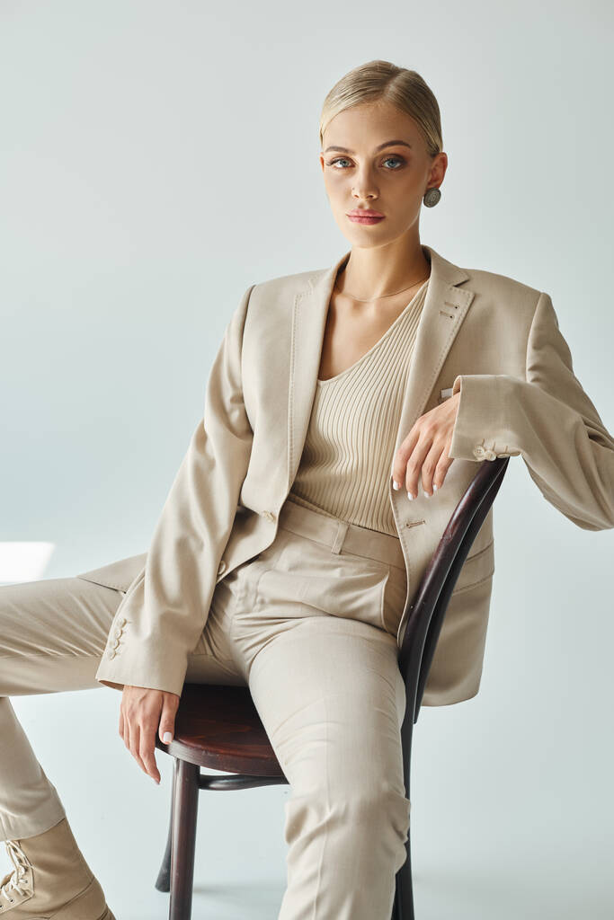 blond mode model in pastel beige pak zitten op stoel en kijken naar de camera op grijze achtergrond - Foto, afbeelding