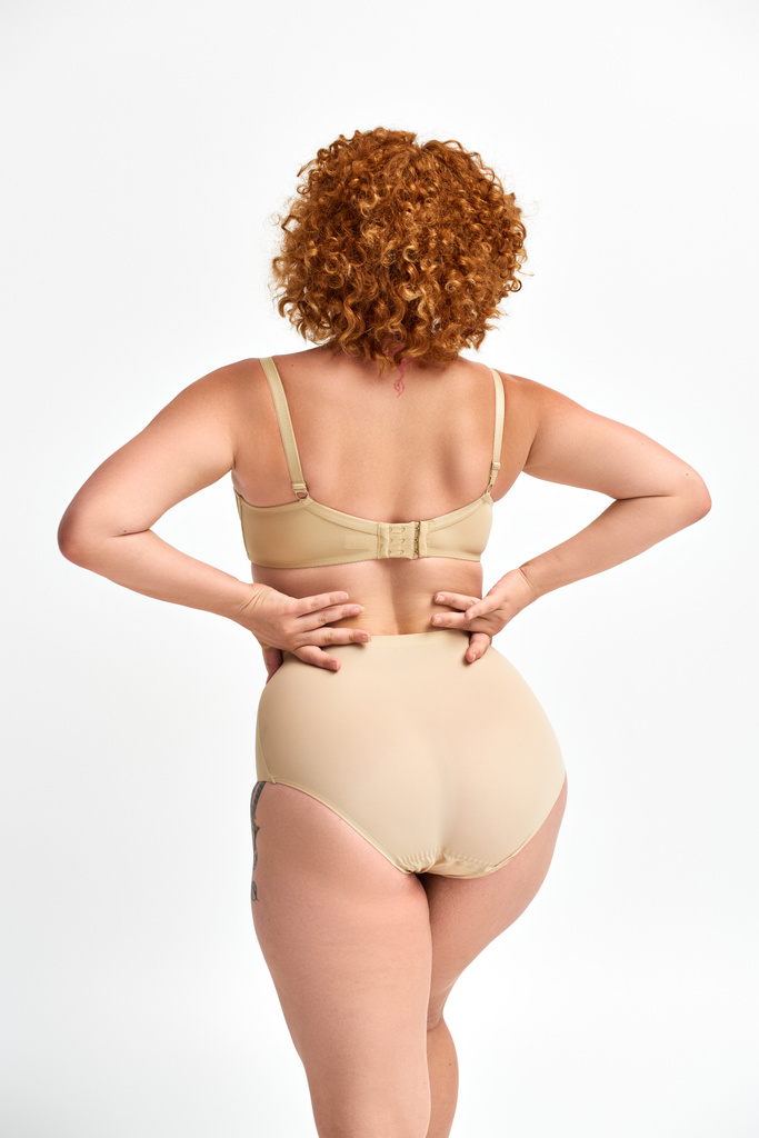 plus size woman with red wavy hair posing in beige lingerie with hands on waist on white, back view - Photo, Image