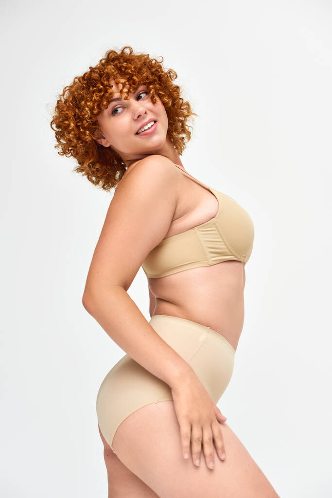 joyful curvy woman in lingerie with red curly hair looking away on white, body positivity - Photo, Image