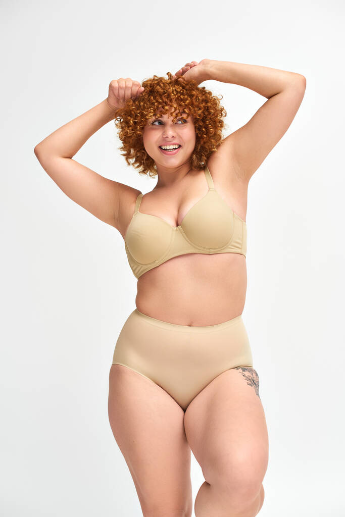 overjoyed and redhead plus size model in beige underwear with raised hands looking away on white - Photo, Image