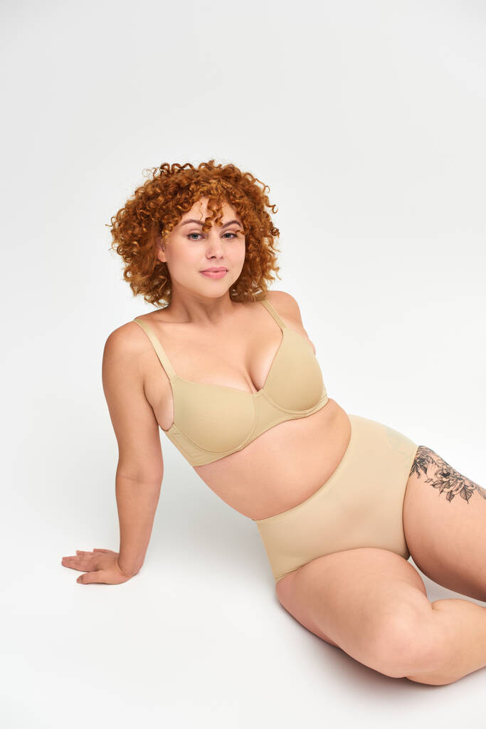 smiling tattooed woman with red wavy hair and plus size body sitting in taupe underwear on white - Photo, Image