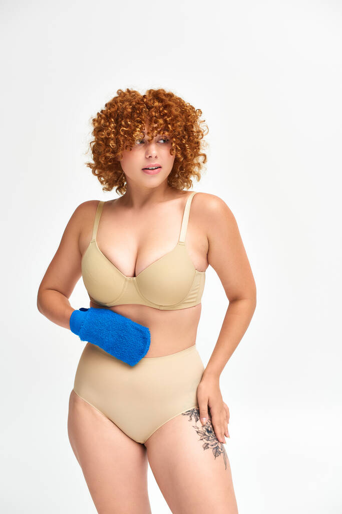redhead plus size woman in beige lingerie holding bath glove and looking away on white - Photo, Image