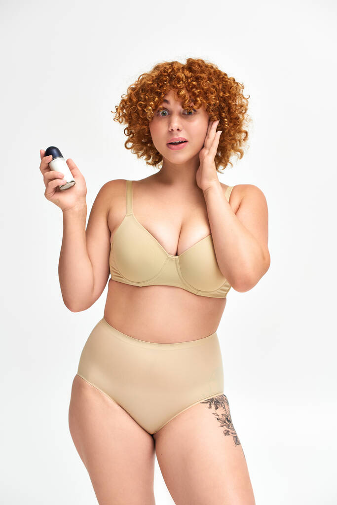 užaslá zrzka plus velikost žena s roll-on deodoranty dotýkající se obličeje a dívá se na kameru na bílém - Fotografie, Obrázek
