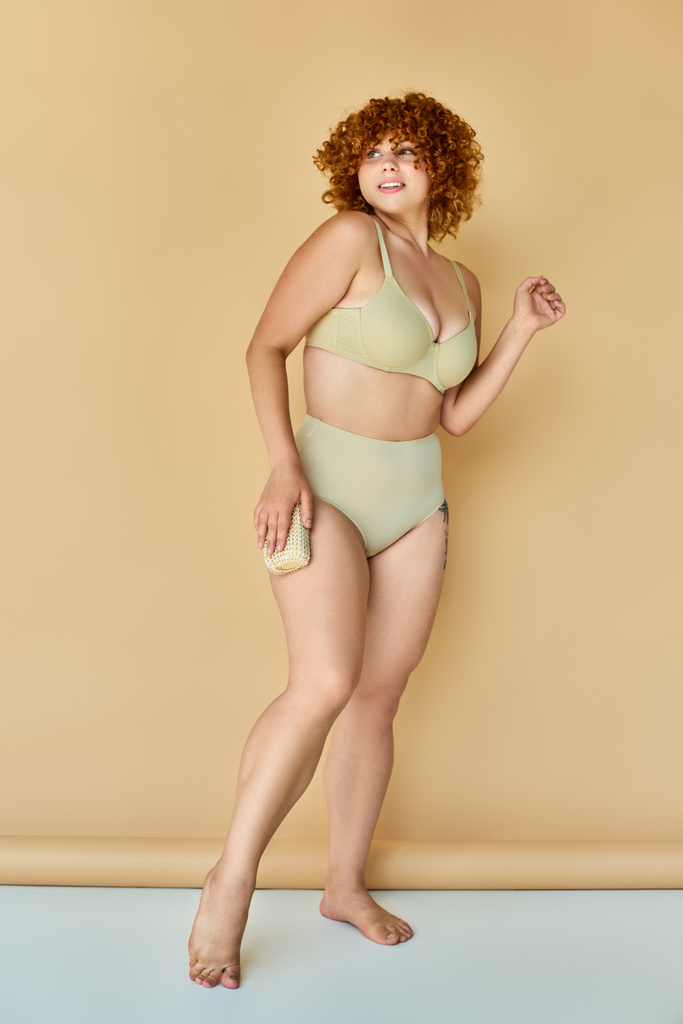 full length of curvy redhead woman in lingerie holding washcloth and looking away on beige backdrop - Photo, Image