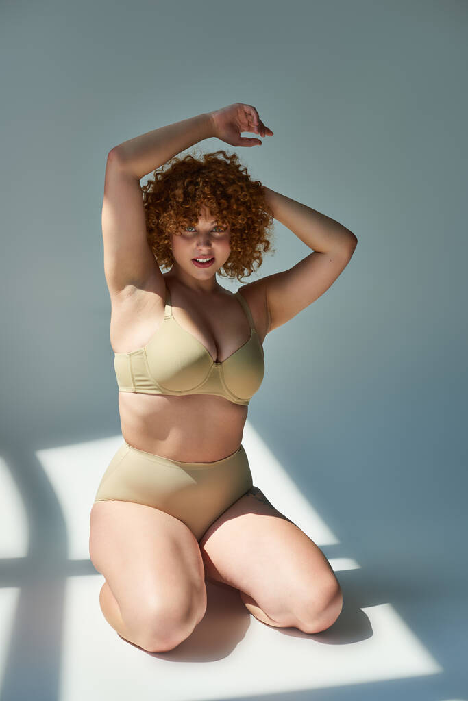 sensual and redhead plus size woman in beige lingerie looking at camera in sunlight on grey - Photo, Image