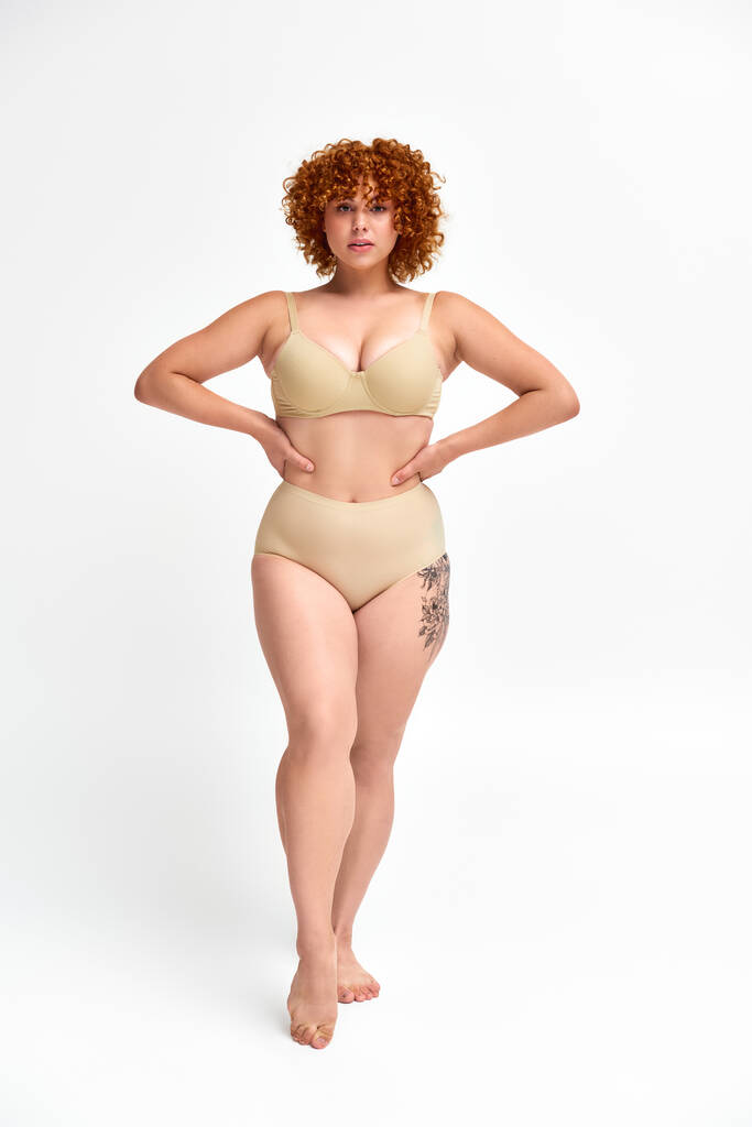 full length of plus size tattooed model in beige lingerie with hands on hips on white backdrop - Photo, Image