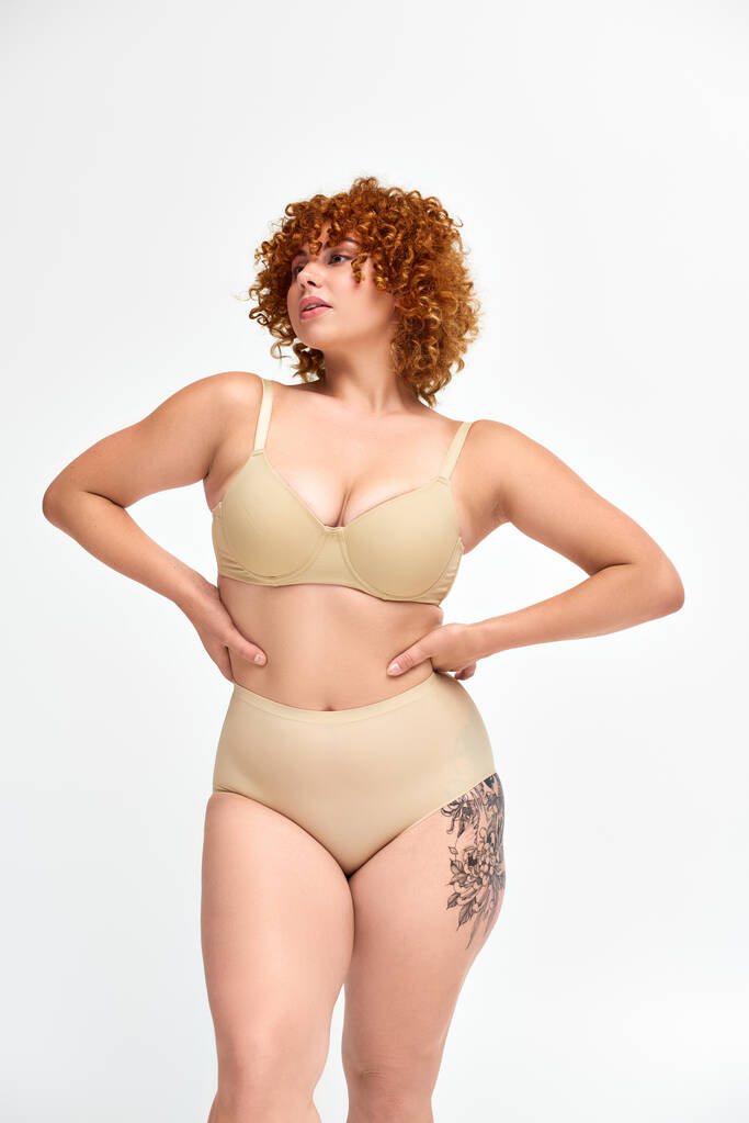 modèle courbé et roux tatoué en lingerie beige avec les mains sur les hanches regardant loin sur le blanc - Photo, image