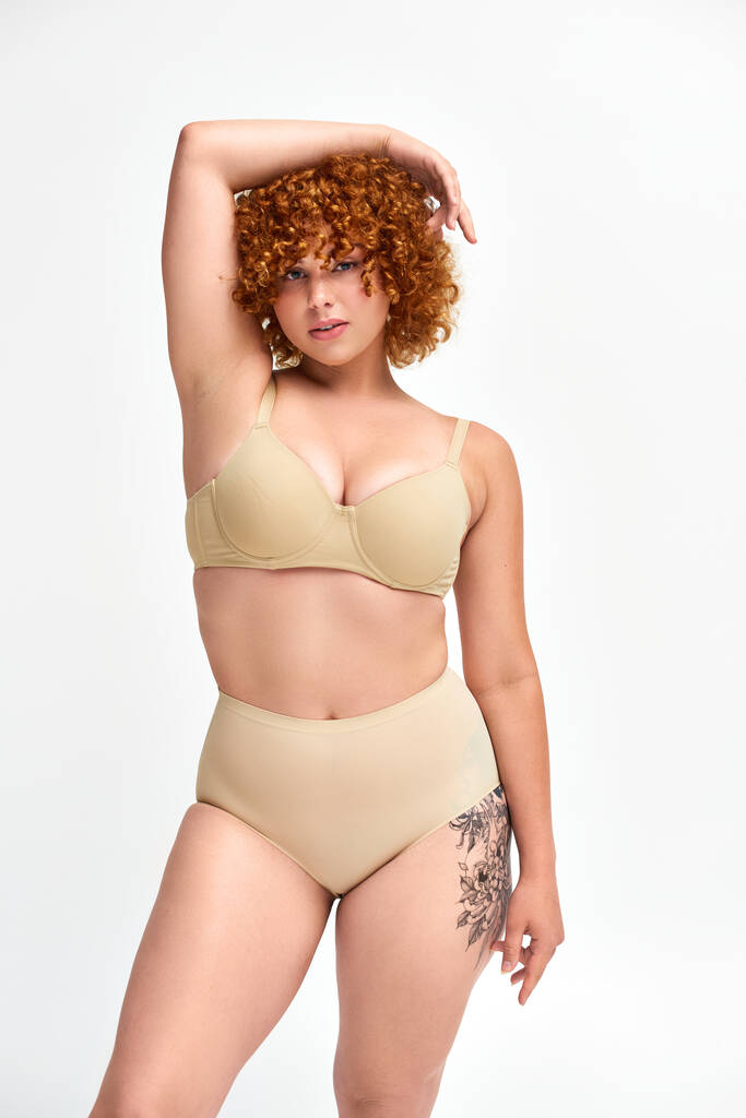 sensuele plus size getatoeëerd model in beige lingerie met handen boven het hoofd kijkend naar camera op wit - Foto, afbeelding