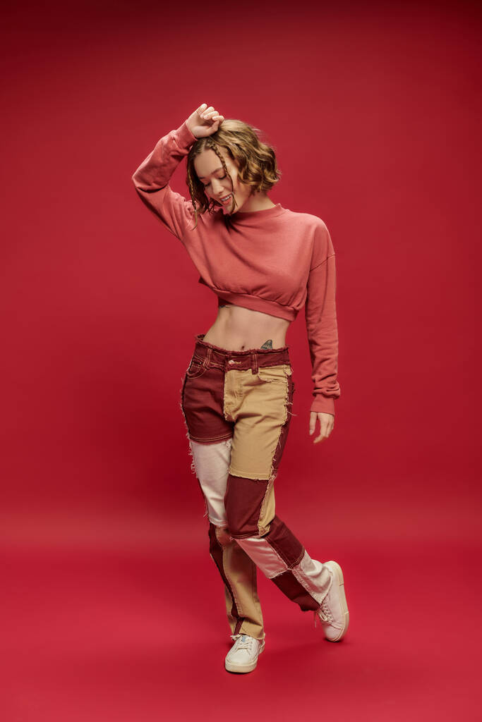 glücklich stylische und junge Frau in Patchworkhosen und abgeschnittenem Langarm posiert auf rotem Hintergrund - Foto, Bild