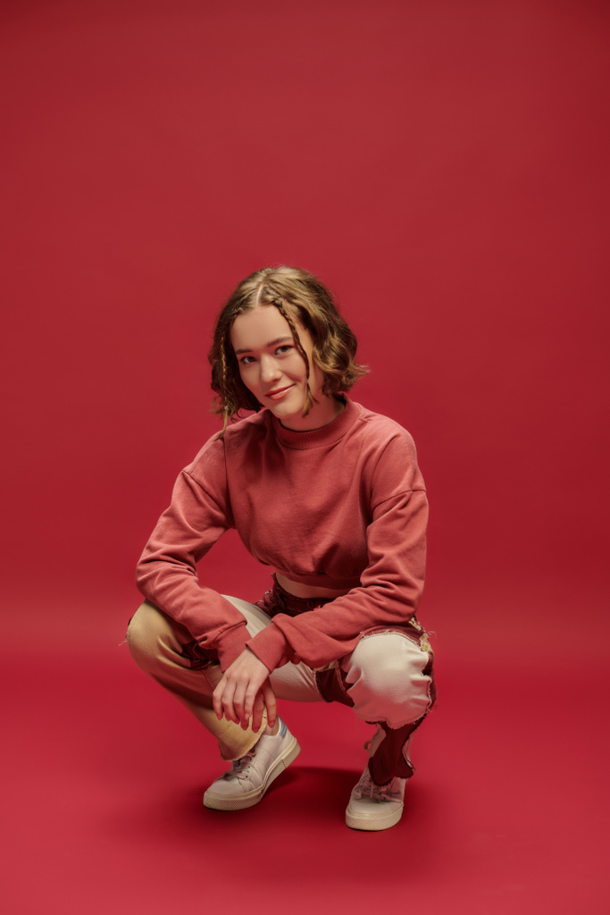 προσωπικό στυλ, νεαρή γυναίκα σε patchwork παντελόνι και περικοπεί μακρύ μανίκι κάθεται σε κόκκινο φόντο - Φωτογραφία, εικόνα