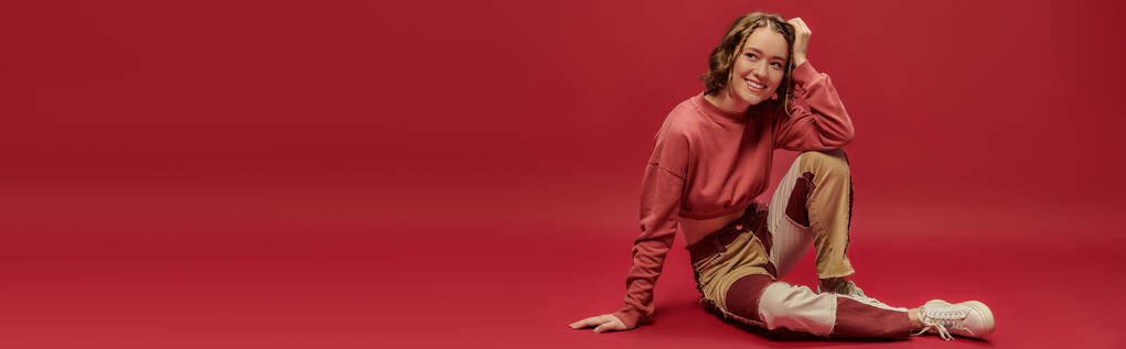 estandarte de mujer alegre en pantalones patchwork y manga larga recortada sentado sobre fondo rojo - Foto, Imagen