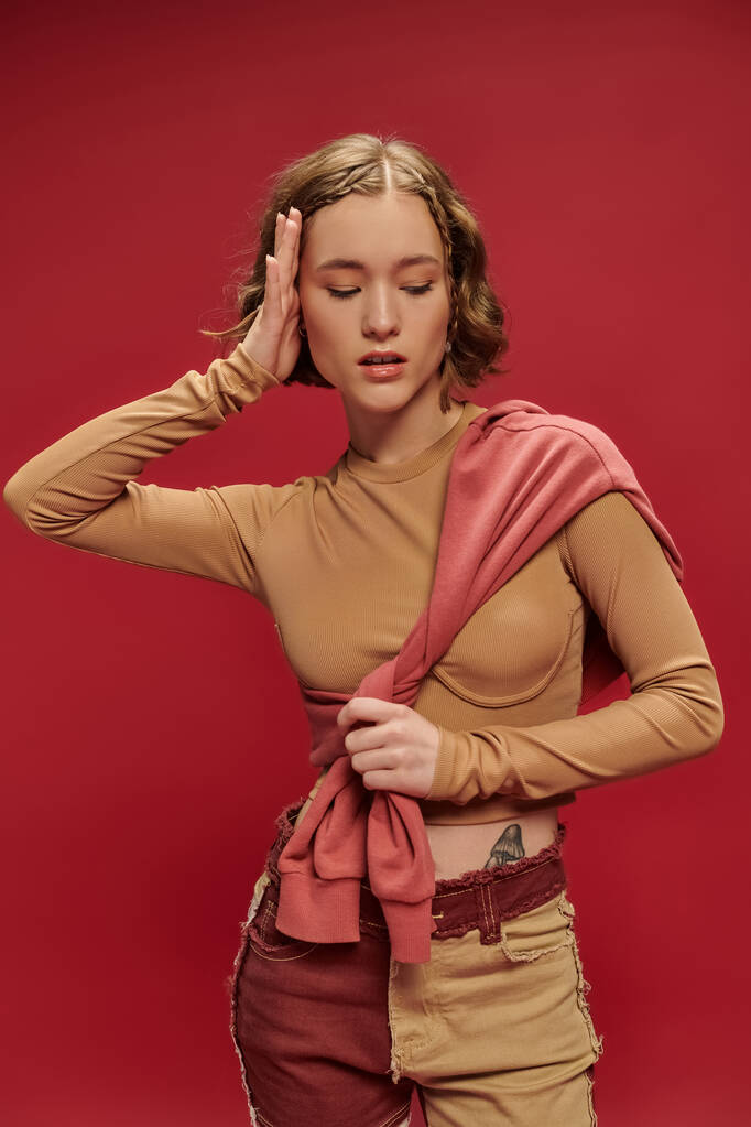 молодая стильная женщина в лоскутных штанах позирует в вязаном свитере на длинном рукаве на красном фоне - Фото, изображение