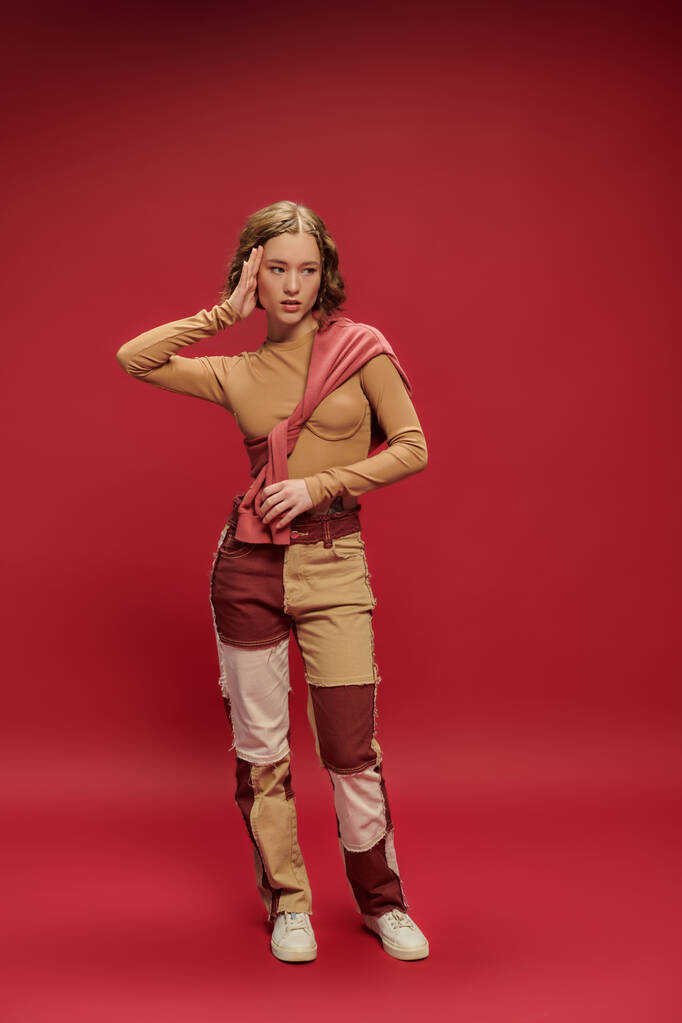 nuori trendikäs nainen tilkkutäkki housut poseeraa sidottu hyppääjä yli pitkähihainen punainen tausta - Valokuva, kuva