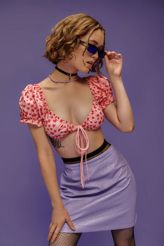 стильная молодая женщина в модном наряде, регулируя солнцезащитные очки на фиолетовом фоне, индивидуальный стиль - Фото, изображение