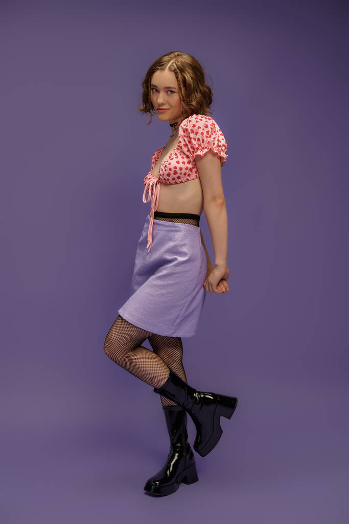 stylish young woman in cropped top and skirt standing in boots on purple background, youth fashion - Photo, Image