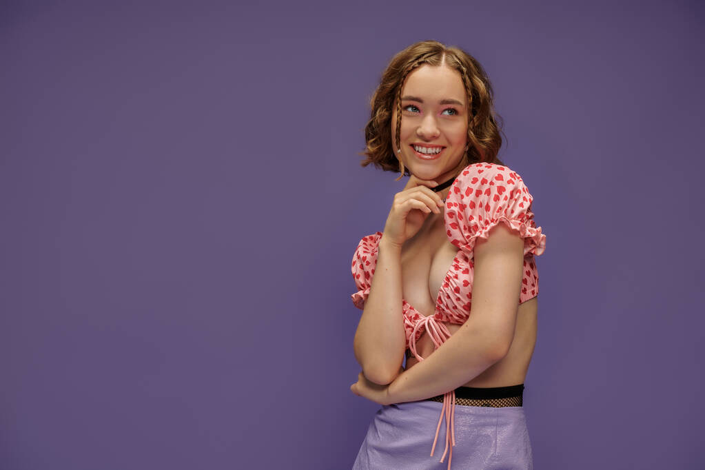 lächelnde junge Frau in abgeschnittenem Top posiert mit der Hand am Kinn auf lila Hintergrund, Emotionen - Foto, Bild