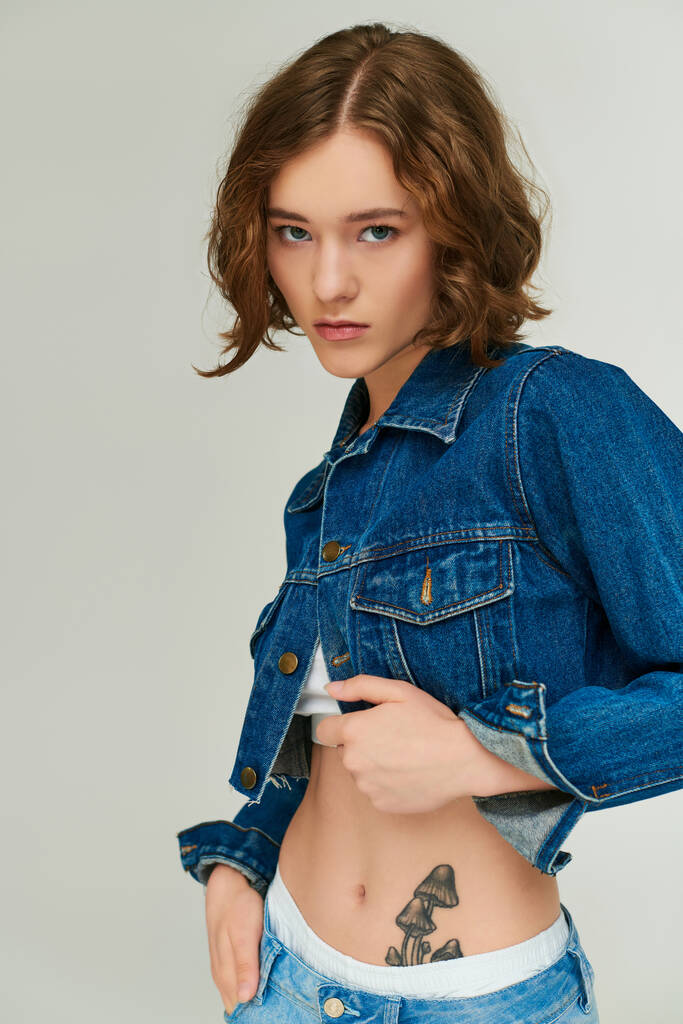 stylowa dziewczyna z tatuażem w przyciętej kurtce jeansowej pozowanie z ręką w kieszeni dżinsów na szarości - Zdjęcie, obraz