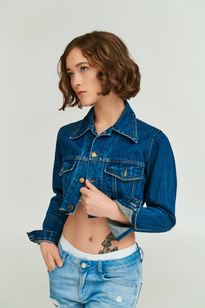 модная девушка с татуировкой в обрезанной джинсовой куртке позирует с рукой в кармане джинсов на сером - Фото, изображение