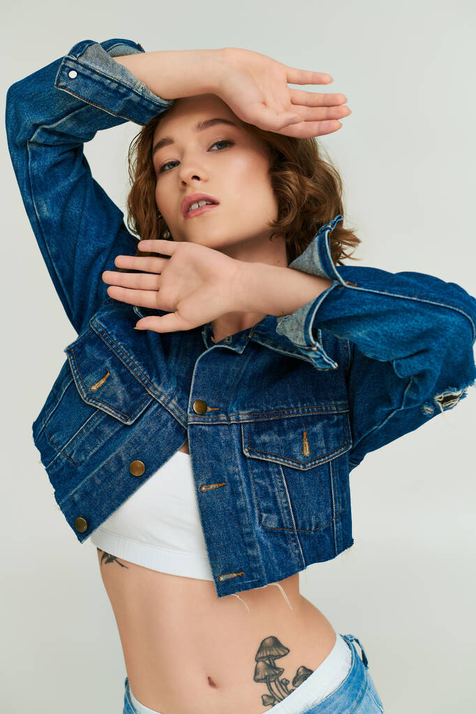 aantrekkelijk jong model met tatoeage in bijgesneden denim jas en blauwe jeans poseren op grijze achtergrond - Foto, afbeelding