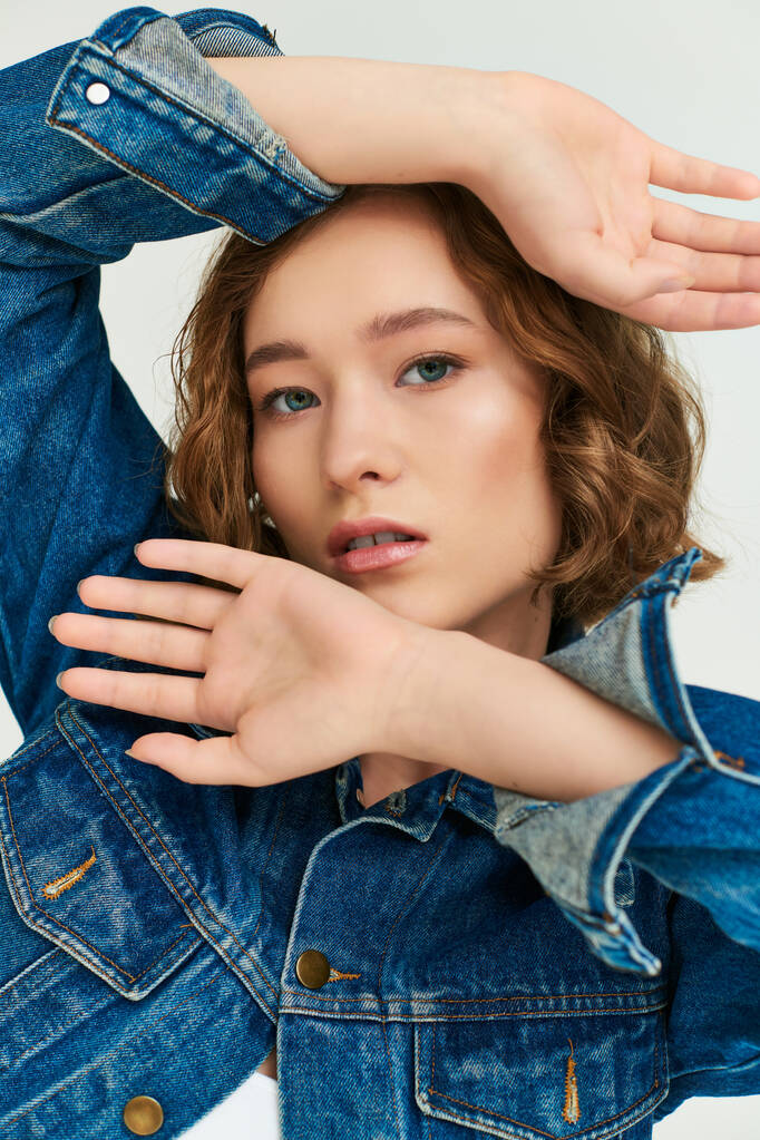 portrait of attractive young model in cropped denim jacket gesturing and posing on grey backdrop - Photo, Image