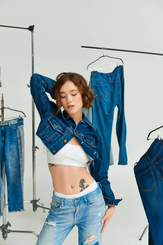 charming young woman with tattoo posing in cropped blue jacket among denim clothes on grey backdrop - Photo, Image