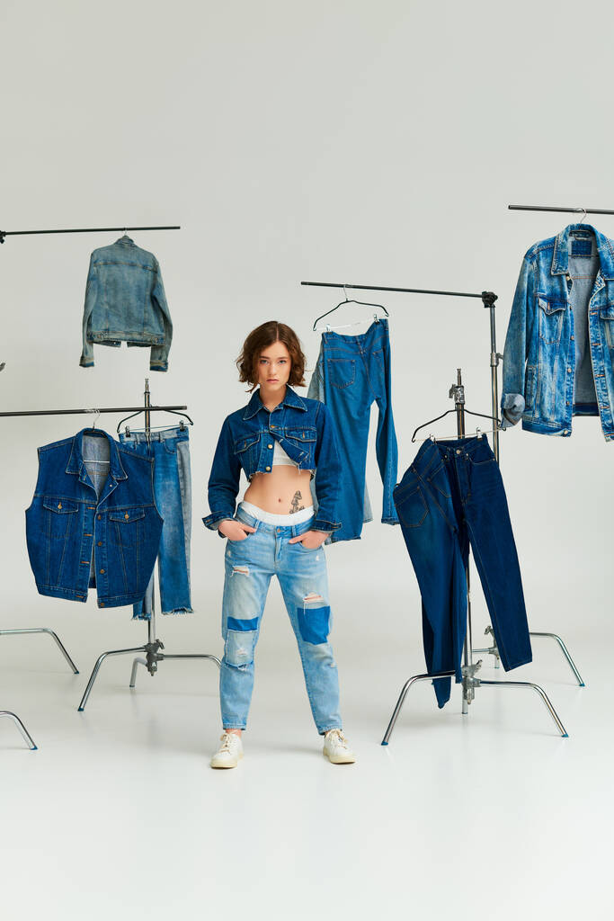 μοντέλο μόδας σε crop σακάκι ποζάρουν με τα χέρια στις τσέπες μεταξύ denim ρούχα σε κρεμάστρες σε γκρι - Φωτογραφία, εικόνα