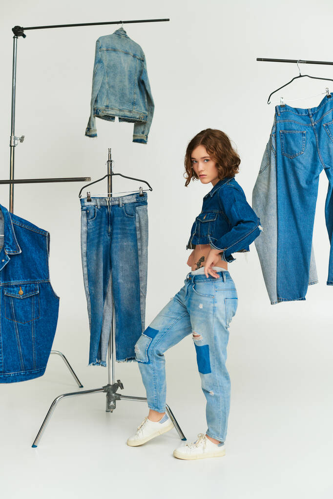 stylisches junges Model mit Tätowierung in abgeschnittener Jacke und Jeans steht zwischen Jeanskleidung auf grau - Foto, Bild