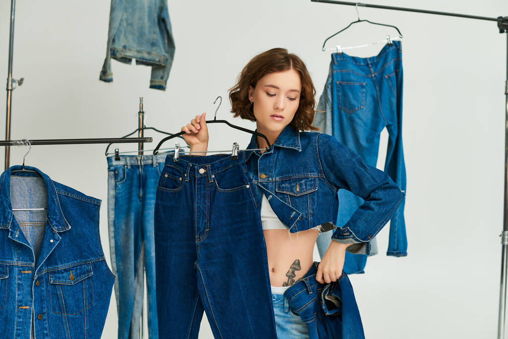 tattooed young woman in blue cropped jacket holding jeans among denim clothes on grey backdrop - Photo, Image