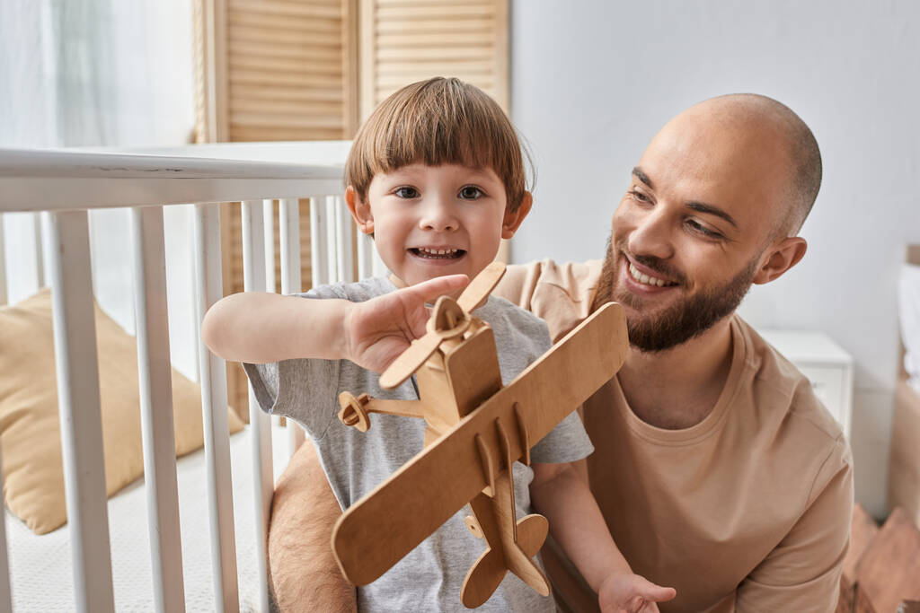 šťastný rozkošný chlapec hraje s dřevěným letadlem se svým otcem a usmívá se na kameru, rodina - Fotografie, Obrázek