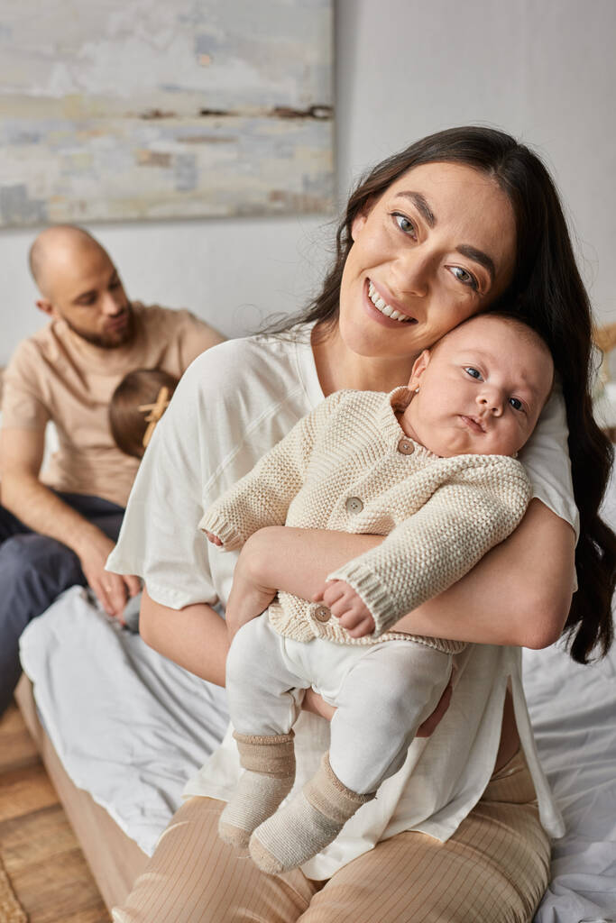 focus op moeder houden pasgeboren baby en glimlachen op de camera met wazig man en zoon op de achtergrond - Foto, afbeelding