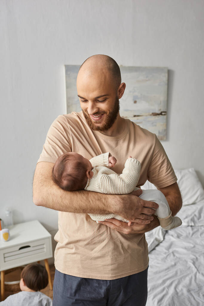 bärtiger Mann in Hauskleidung hält sein neugeborenes Baby in der Hand und lächelt es glücklich an, Familienkonzept - Foto, Bild