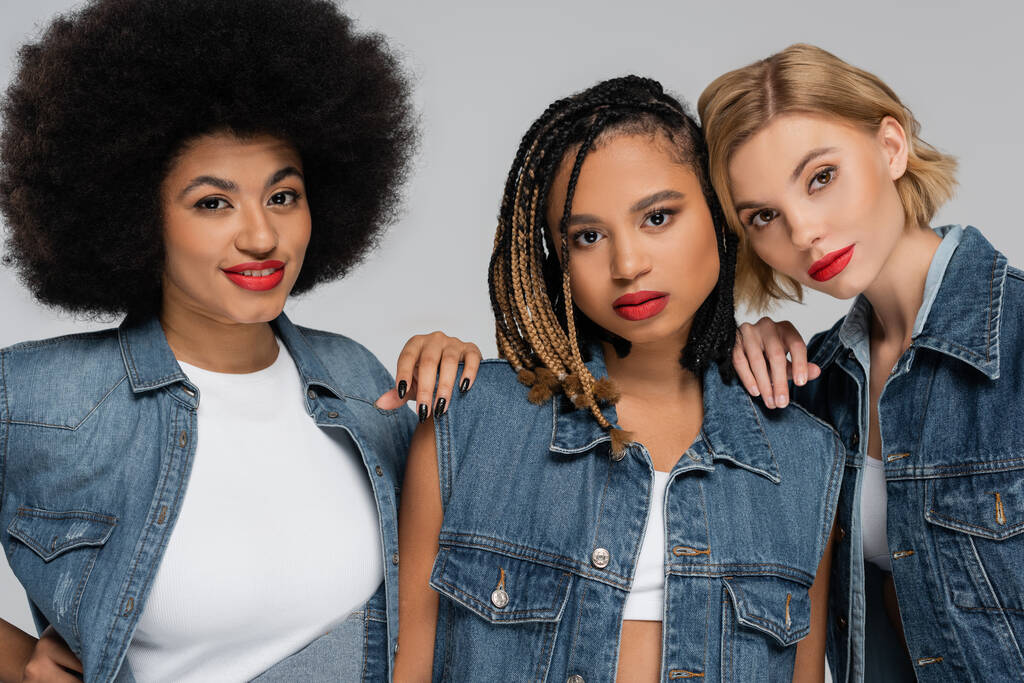 verführerische multiethnische Freundinnen in Blue-Jeans-Klamotten, die in die Kamera blicken, auf grau, Streetwear - Foto, Bild
