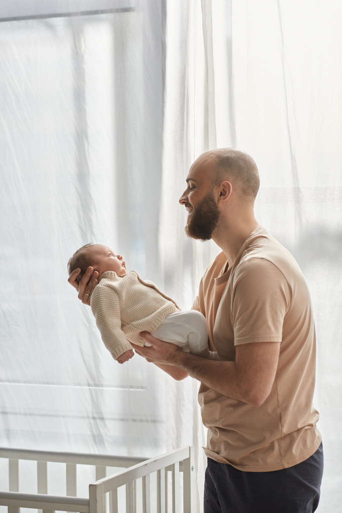 вертикальный снимок счастливого отца в уютной домашней одежде, держащего своего новорожденного сына и с любовью улыбающегося ему - Фото, изображение