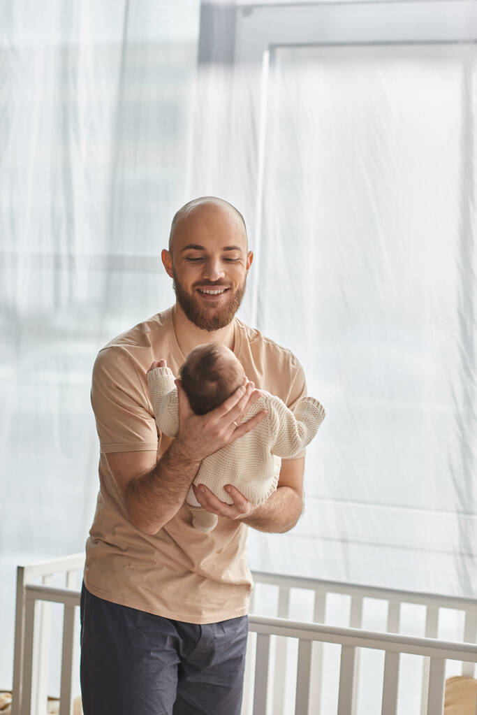 tiro vertical de padre barbudo bien parecido sosteniendo a su bebé junto a la cuna, concepto de familia - Foto, Imagen