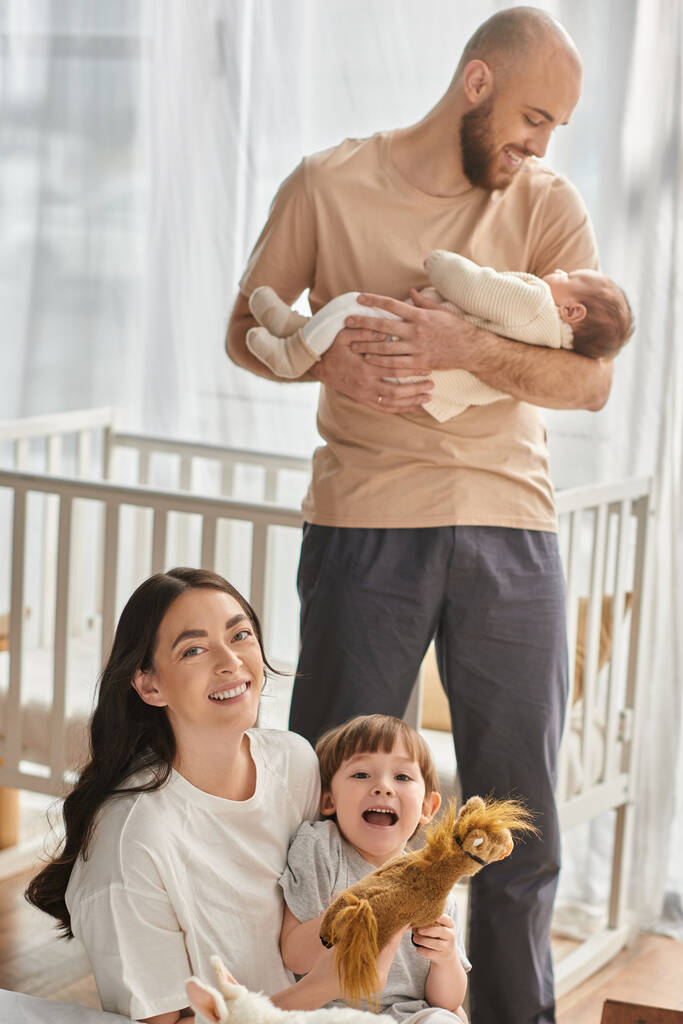 centrarse en la madre jugando con su hijo y sonriendo a la cámara junto a su marido borroso sosteniendo al bebé - Foto, Imagen